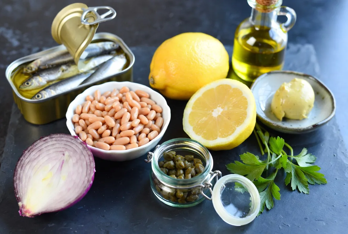 Fresh ingredients for baked sardine and white bean salad including capers, flat-leaf parsley, lemon, and olive oil on a kitchen surface