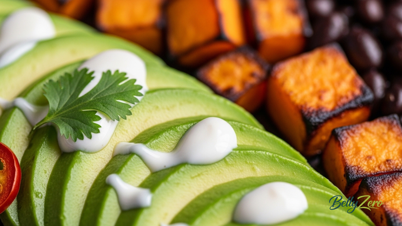 Close-up of avocado slices with white lime crema drizzle and caramelized sweet potato cubes in the black bean sweet potato bowl. BellyZero.